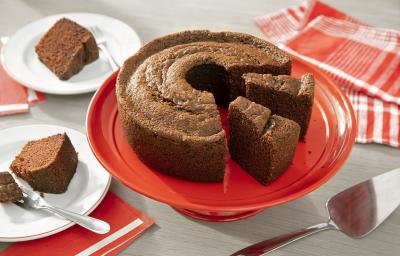 Fotografia em tons de marrom em uma bancada de madeira de cor cinza. Ao centro, uma boleira vermelha contendo o bolo de chocolate. Ao fundo, há um pano vermelho listrado e ao lado, há 2 pires com 2 fatias de bolo.