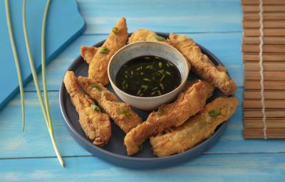 Foto da receita de Costelinha de Tilápia Copacol Empanada com Molho Oriental na Airfryer, servida em um tigela azul escura, com um pote pequeno com o molho no centro do prato, tudo numa bancada azul