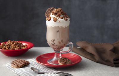 Foto de uma mesa cinza. Ao centro há um prato vermelho com uma caneca alta com a bebida de Chocolate Quente, decorada com Chantilly e o crumble. Na base há um biscoito Calipso e mais dois na mesa. Ao fundo há um prato com mais crumble.