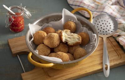 Foto em tons de marrom da receita de bolinho de mandioca (aipim) com queijo servida em diversas porções em um escorredor sobre uma tábua de madeira em cima de uma mesa cinza chumbo. Ao lado há uma escumadeira prateada e um paninho colorido