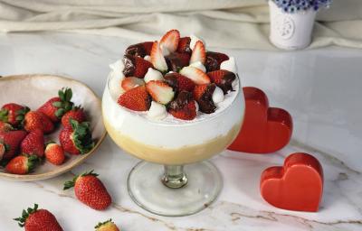 Foto da receita de Morangoffee na Taça, servida em uma grande taça de vidro, em duas camadas, uma de creme bege e outra de creme branco, decorada com morangos com chocolate e suspiros, em uma bancada com mais morangos e alguns corações decorativos