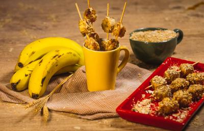 Foto da receita de Banana e Neston no espeto, com alguns espetos dentro de uma caneca amarela e outros em um prato, tudo em uma bancada de madeira decorada com bananas, trigo e um caneca com Neston 3 Cereais dentro.