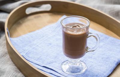 Fotografia em tons de azul e marrom, com bandeja ao centro sobre toalha cinza, dentro um copo com chocolate quente sobre guardanapo azul.