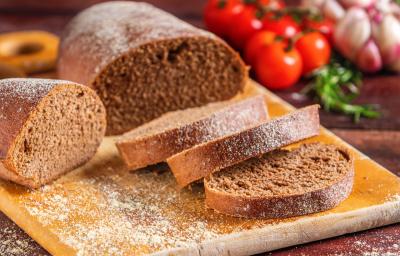 Fotografia de uma tábua média de madeira sobre uma mesa de madeira escura. Sobre a tábua tem uma receita de pão australiano com três fatias cortadas e sobrepostas ao lado de dois pedaços maiores do pão. Fotografia de uma tábua média de madeira sobre uma mesa de madeira escura. Sobre a tábua tem uma receita de pão australiano com três fatias cortadas e sobrepostas ao lado de dois pedaços maiores do pão.