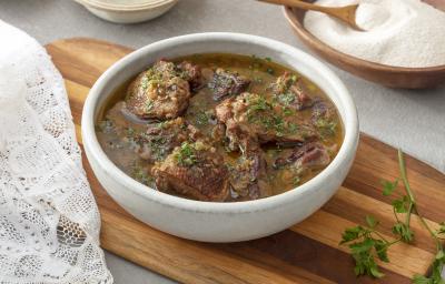 Foto em tons de marrom da receita de pato no tucupi servida em um bowl de cerâmica branco sobre uma tábua de madeira com cheiro verde ao lado. Ao fundo há um recipiente de madeira com farinha de mandioca e ao lado um paninho branco