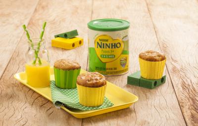 Fotografia em tons de amarelo e verde em uma bancada de madeira clara, um recipiente amarelo com dois cupcakes de maçã e aveia em forminhas amarela e verde. Ao lado, brinquedinhos infantis, uma lata de Ninho Fases e mais um cupacke.