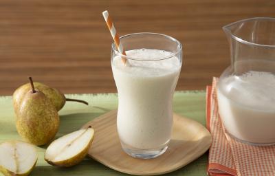 Fotografia em tons de branco em uma bancada de madeira de cor verde, ao centro, um pires de madeira contendo um copo com o smoothie. Ao lado, há algumas peras e do outro lado, um pano laranja quadriculado com uma jarra em cima.