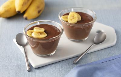 Fotografia em tons de azul e marrom de uma bancada com um paninho azul, duas tacinhas transparentes com o creme de chocolate e rodelas de banana decorando, ao lado duas colheres pratas e ao fundo 3 bananas.