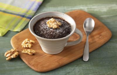 Fotografia em tons de verde em uma bancada de madeira verde, uma tábua de madeira, uma caneca bege com o brownie de chocolate com nozes dentro dela. Ao lado, nozes caídas e um paninho verde.