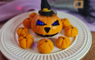 Fotografia em tons de laranja, em uma bancada de madeira, um prato branco ao centro com os beijinhos em formato de abóbora. No centro, uma abóbora pequena com desenho de Halloween e um chapeuzinho de bruxa ao fundo da foto.