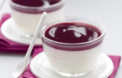 Fotografia em tons de roxo escuro em uma bancada de madeira cinza, dois guardanapos de pano roxos, dois pratinhos de café brancos e dois copinhos de vidro com a sobremesa pannacotta com calda de vinho tinto dentro.