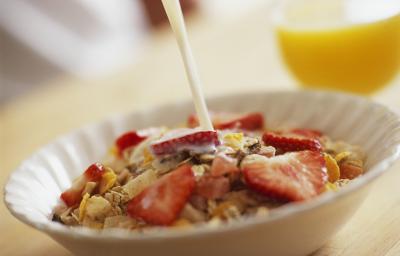 Fotografia de um recipiente branco de vidro fundo um creme com frutas e cereal Nesfit. O recipiente está sobre uma mesa de madeira, e ao fundo tem um copo com suco de laranja.