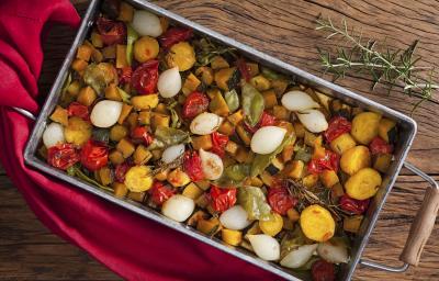 Fotografia de um recipiente de ferro com alças de madeira, dentro estão todos os legumes ficados em cubos assados, e ao lado está um guardanapo de tecido vermelho.