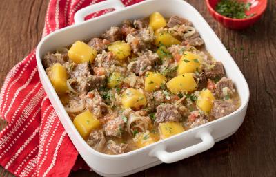 Fotografia em tons de marrom e branco uma bancada vista de cima, ao centro um recipiente quadrado com alças cor branco, dentro dele cubos de carne e pedaços de Mandioquinha e Quinoa. Ao lado um pano listrado na cor vermelho e branco.