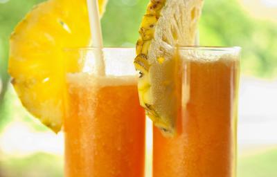 Fotografia de dois copos de vidro grandes com suco de laranja, abacaxi e mel. Em cada copo, tem metade de uma fatia de abacaxi e tem também um canudo. Ao fundo tem uma paisagem com de natureza com árvores.