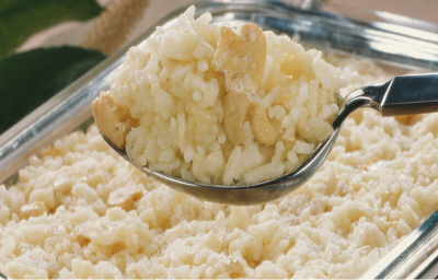 Fotografia em tons de branco e bege, ao centro colher de servir com arroz e cogumelos sobre refratário com o mesmo arroz, ao fundo folhas verdes como decoração.