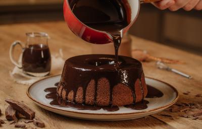 Fotografia em tons de vermelho em uma mesa de madeira com um prato branco com o bolo de chocolate em cima e uma calda de chocolate por cima. Uma panela vermelha em cima do bolo derramando a calda de chocolate.