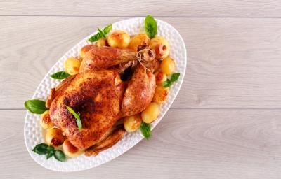 fotografia em tons de cinza, branco e bege de uma bancada cinza vista de cima. Contém um recipiente oval branco com frango assado e batatas bolinhas ao redor.