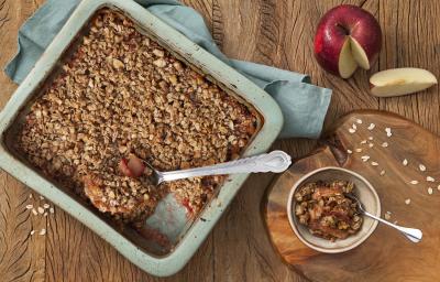 Fotografia em tons de azul em uma mesa de madeira escura com uma travessa retangular azul e o crumble de maçã e aveia dentro. Ao lado, uma tábua de madeira e um potinho com um pedaço do crumble. Ao fundo, uma maçã cortada ao meio.