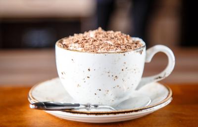 fotografia em tons de branco e marrom de uma bancada marrom vista de frente. Ao centro contém um pratinho redondo branco com uma xicara branca por cima, dentro da xicara contém a bebida e por cima raspas de chocolate, ao lado uma colher para mexer.