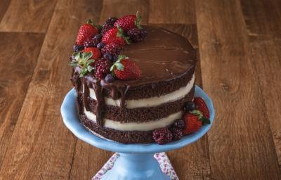 Imagem da receita de Naked Cake de Cacau, decorado em camadas marrons e brancas, com chocolate escorrendo, decorado com frutas vermelhas. O bolo está sobre um prato azul em uma bancada de madeira.