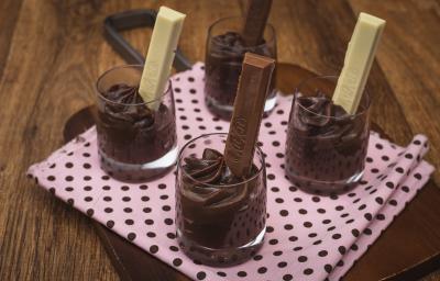 Fotografia em tons de rosa em uma bancada de madeira, uma tábua de madeira com um pano rosa e bolinhas pretas, quatro copinhos de vidro com brigadeiro de colher de chocolate dentro de cada um com um KitKat ao Leite e KitKat Branco dentro dos copinhos.