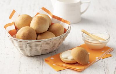 Fotografia em tons de laranja e branco de uma bancada branca com um paninho laranja, sobre ele um pãozinho aberto recheado e um recipiente de vidro com requeijão. Ao lado uma cesta com um paninho laranja e branco com os pães.