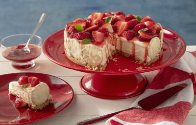 Fotografia em tons de vermelho em uma bancada de madeira de cor branca. Ao centro, uma boleira vermelha contendo a torta holandesa. Ao lado, há um pires com uma fatia da torta e um pano listrado vermelho.