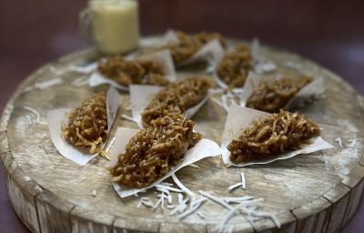 Foto da receita de cocada com doce de leite em tons de marrom servidas em oito porções em lenços de papel sobre uma tábua de maderia redonda e grossa. Ao fundo, há um recipiente de vidro com leite MOÇA para decorar