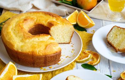 Fotografia de um bolo de laranja feito em forma de furo central sobre um prato raso de cerâmica branco. Ao lado tem um prato de sobremesa com uma fatia do bolo e um garfo, e ao redor tem pedaços e fatias de laranja, sobre uma mesa de madeira branca.