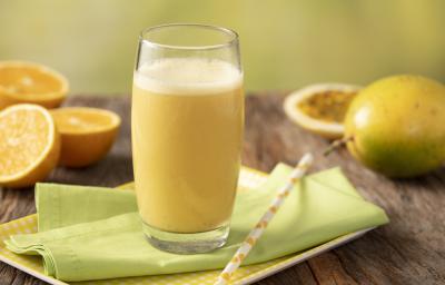 Fotografia em tons de amarelo, verde e marrom de uma bancada de madeira com uma bandeja amarela e um paninho verde com um canudo sobre ele um copo de vidro com a vitamina de laranja. Ao fundo três metades de laranjas, um maracujá inteiro e uma metade.