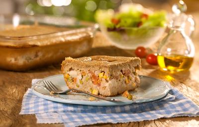 Fotografia em tons de marrom em uma bancada de madeira com um pano xadrez azul e um prato branco com um pedaço de torta integral de legumes. Ao fundo, uma travessa com a torta inteira, um potinho com salada e um vidro de azeite.