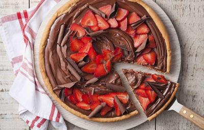 Fotografia em tons de branco e vermelho de uma bancada de madeira branca com uma tabua redonda com a pizza de chocolate com morangos. Ao lado um paninho branco com listras vermelhas e uma espátula retirando uma fatia da pizza.