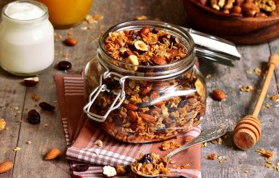 Fotografia em tons de cinza com um pote de vidro ao centro. Dentro do pote existe uma granola caseira feita com castanhas, aveia, passas e sementes. Ao lado existe um pote de iogurte.