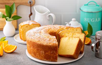 Fotografia de um bolo de laranja feito em forma redonda com furo no meio, o bolo está sobre um apoio, e tem três fatias cortadas, uma apoiada na outra. Ao fundo tem uma fatia do bolo em um prato de vidro pequeno, ao lado de pedaços de laranja.