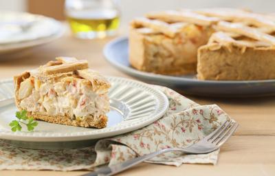 Fotografia em tons de bege em uma mesa de madeira com um pano florido em bege e vermelho e um prato branco apoiado nele com um pedaço de torta de palmito e alho-poró. Ao fundo, um prato grande com a torta cortada ao meio.