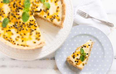 fotografia em tons de branco e amarelo de uma bancada branca vista de cima. Contém um prato branco redondo com uma torta com mousse de maracujá e sementes para decorar, e ao lado um prato redondo azul com a fatia da torta.