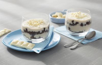 Fotografia em tons de azul em uma bancada de madeira cinza, um pano azul, um prato redondo azul claro e duas taças de vidro com o pavê de chocolate ao leite com chocolate branco, Galak. Ao fundo, um potinho com quadradinhos de chocolate Galak.