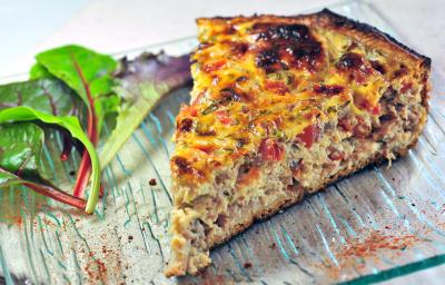 Fotografia mostra uma fatia de torta com camadas bem definidas de massa leve e recheio colorido de sardinha, tomate e azeitonas, polvilhada com orégano.