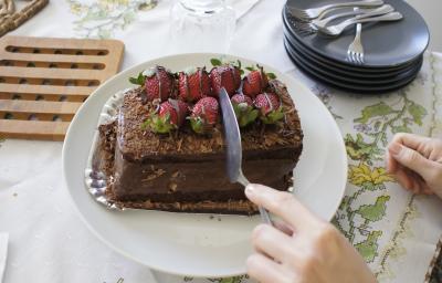 Fotografia de uma mão cortando um bolo de chocolate recheado com morango que está em um prato branco. Ao fundo, alguns pratos pretos empilhados, e ao lado, uma decoração em madeira para apoiar bolo ou pães. Tudo está sobre uma toalha de mesa branca.