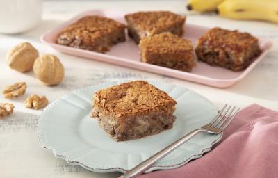 Foto da Receita de Bolo de Banana, Leite Moça e Nozes Caramelizadas. Observa-se uma fatia de foto no plano principal com um prato de sobremesa azul e decorados com banana e nozes.