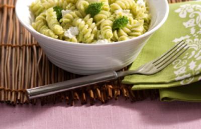 Fotografia em tons de verde em uma mesa de madeira com uma toalha rosa e um jogo americano de palha marrom.  Ao centro, um prato branco fundo redondo com o macarrão tipo parafuso com o molho pesto de brócolis e ao lado um guardanapo de pano verde.