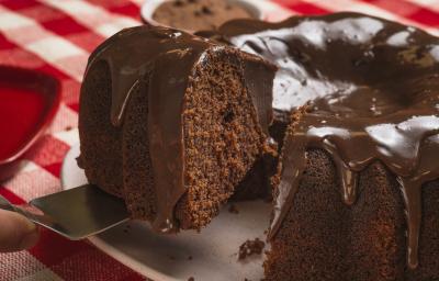 Foto bem aproximada de um bolo de chocolate com uma fatia sendo retirada. O bolo está num prato branco e coberto com Brigadeiro e o prato está sobre uma toalha xadrez vermelho e branco.