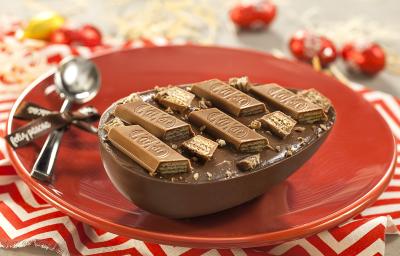 Fotografia em tons de vermelho em uma bancada de madeira cinza, um pano vermelho com listras brancas, um prato vermelho redondo raso com um ovo de Páscoa recheado com ganache e KitKat de Chocolate ao Leite.