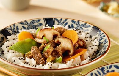 fotografia em tons de branco de bege de uma bancada bege vista de frente. Contém dois recipientes redondos em tons de branco e azul com arroz, cenouras fatiadas e shitake, ao lado o hashi para servir e ao fundo uma xicara branca.