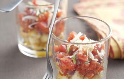 Fotografia em tons de amarelo e vermelho em uma bancada cinza com uma toalha com detalhes amarelos e vermelho. Ao centro, dois copinhos de vidro com a bruschetta com tomate, croutons e queijo dentro deles.