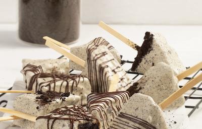 Fotografia em tons de branco em uma bancada de cor branca. No centro, estão os Cakes Pop Cookies’n Cream polvilhados com chocolate. Ao fundo, um recipiente contendo Topping de Negresco.