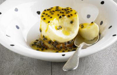 Fotografia de um flan de maracujá e manga, com calda de maracujá em cima e no fundo do prato, que tem cor branca com bolinhas pretas. Apoiado no prato, uma colher de sopa segurando um pedaço do flan. A sobremesa está sobre uma mesa de madeira cinza.