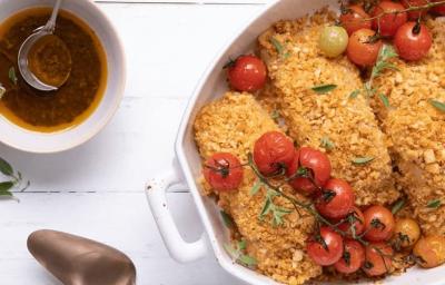 Fotografia em tons de amarelo em uma bancada de madeira branca com um refratário oval branco com as postas de bacalhau com crosta de pão e tomatinhos cereja assados dentro dele.