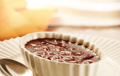 fotografia em tons de branco e marrom de uma bancada vista de frente, contém um prato redondo e branco com um potinho oval branco com um creme de chocolate dentro e uma colher ao lado para servir.